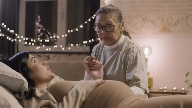 Asian doula holding pregnant womans hand and talking with her. Expecting mother lying on bed, stroking big belly, taking care of future baby. Motherhood, pregnancy, childbirth concept