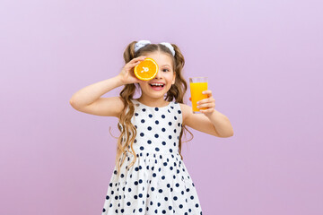 A little girl holds orange juice and an orange. A child in a polka dot dress drinks freshly...
