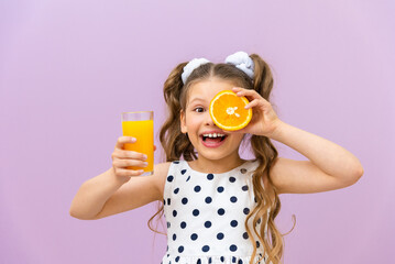 A little girl holds orange juice and an orange. A child in a polka dot dress drinks freshly...