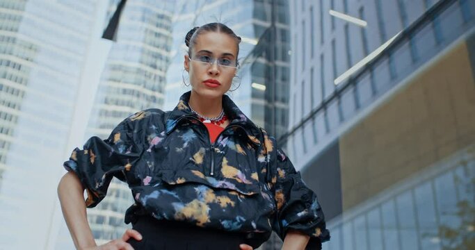 Urban style seductive girl wearing colored jacket and cool futuristic glasses posing with hands on hips looking at camera standing near high skyscrapers modern buildings. Handsome female fashion model