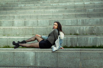 Fototapeta na wymiar A beautiful girl in a black skirt and denim jacket poses sitting on the steps