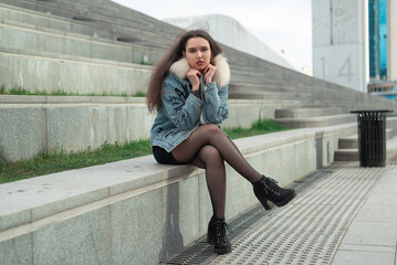 A beautiful girl in a black skirt and denim jacket poses sitting on the steps