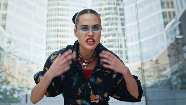 Urban style cheeky female wearing colored windbreaker and cool futuristic glasses rapping aggressively while sitting on stairs in downtown overlooking high-rise skyscrapers. Handsome fashion model