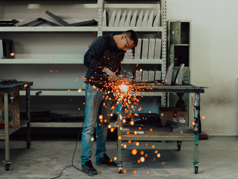 Heavy Industry Engineering Factory Interior with Industrial Worker Using Angle Grinder and Cutting a Metal Tube. Contractor in Safety Uniform and Hard Hat Manufacturing Metal Structures.