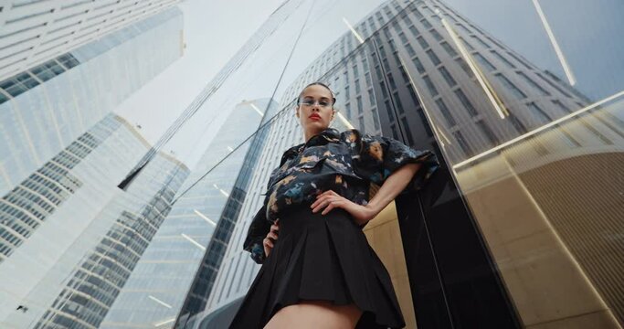 Wide angle urban style seductive girl wearing short skirt colored jacket cool futuristic glasses looks into camera standing by glass wall high skyscraper modern building. Handsome female fashion model