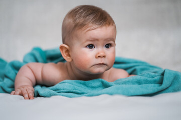 cute little naked 6 month old baby lying on stomach and looking up in prone position after bathing wrapped in turquoise towel at home