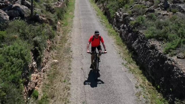 Middle age man wearing a cap, sunglasses and an orange shirt riding a BTT following an old railway path