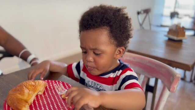 Exotic and cute afro european two year old child wearing a  eating a croissant and playing with a plastic fork, unfocused background