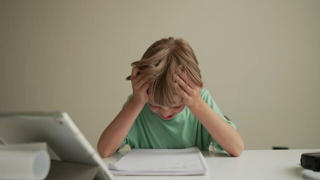 7 yeas old boy sitting at table in room and crying while working on hard homework. Frustrated young boy lays head done on schoolwork.