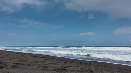 Long waves of surf roll onto the beach. Foam on black volcanic sand. Blue sky with clouds. Kamchatka. Khalaktyrsky beach