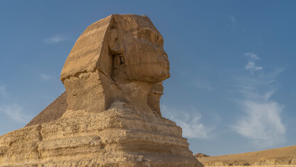 A fragment of the sculpture of the Great Sphinx- the head and part of the body. Close-up. Profile view. The texture of sandstone is visible. Background- blue sky, clouds. Egypt. Giza