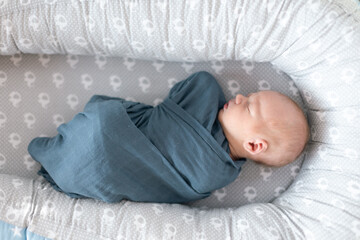 Newborn baby boy sleeping and swaddled in blue cloth lying in grey nest.