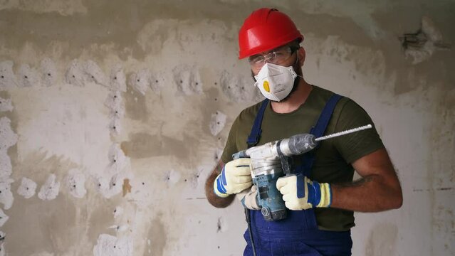 Contractor standing, raising rotary hammer drill in hands in apartment renovation interior. Construction worker looking at camera for low angle hero shot. Man in protective glasses, hardhat, overalls.