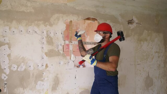 Contractor destroys wall with a sledgehammer making hole for rearrangement. Man doing manual dismantling and demolition works with big hammer hits for apartment renovation. Construction worker has fun