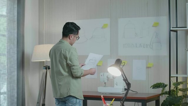 Back View Of Asian Man Designer Compare The Bag Design Sketch Paper In Hand To The Paper On The Wall
