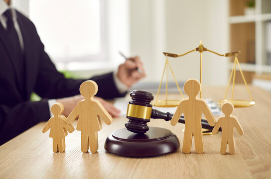 Gavel, sound block and little wooden figures of parents and children placed on desk in courthouse up close, judge and scales of justice in background. Family law, court, divorce, child custody concept