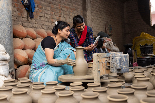 Mother and daughter using digital tablet while making clay pot on spinning wheel at village