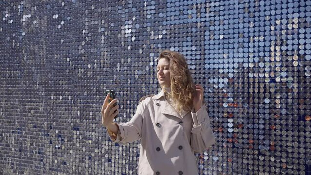 Beautiful woman in beige coat makes selfie at shiny mirror pieces wall during stroll. Young chestnut woman makes photos on sunny day taking city walk