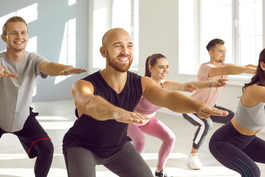 Group of happy, positive, smiling young people having a fitness workout at the gym. Cheerful sporty well trained men and women in comfortable sportswear doing a squats exercise together