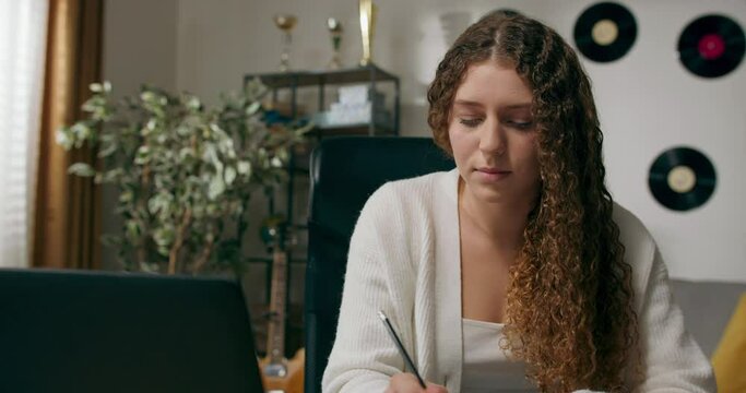 Girl with curly hair sits at desk in living room and studies on laptop taking notes focused young woman work on computer writes in notebook takes online course or training at home concept of education
