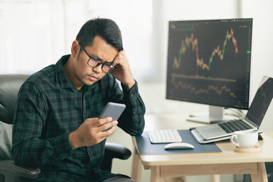 Asian man trader unhappy and serious sitting at home office in front of monitors with cryptocurrency graph holding smartphone monitoring cryptocurrency price