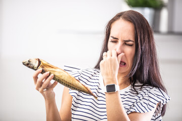 Woman holds stinky fish holds her nose with disgusted facial expression from the smell - obrazy, fototapety, plakaty