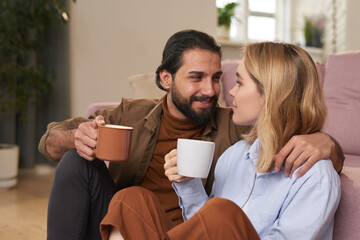 Modern young heterosexual couple sitting on floor close to each other in living room drinking coffee and chatting about something