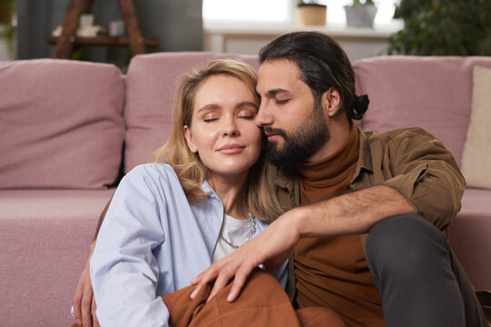 Portrait of lovey-dovey couple sitting on floor against sofa in living room relaxing with eyes closed