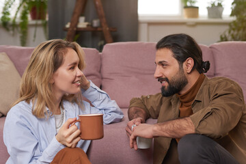 Medium close-up portrait of joyful couple sitting on floor against sofa in living room drinking tea and discussing something