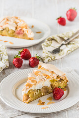 Piece of rhubarb pie with meringue topping and roasted almonds on white wooden background