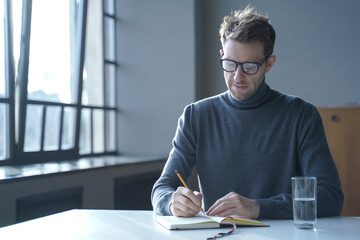 Confident focused German man freelancer in glasses taking notes in agenda sitting at desk at home