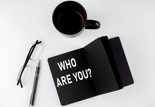 WHO ARE YOU written text in small black notebook with coffee , pen and glasess on white background. Black-white style