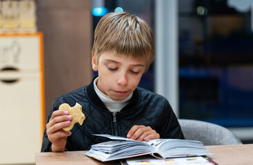 Teen boy with open book in hands and healthy grain cookies reading in cafe at night. Cafe city...
