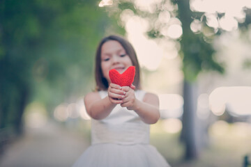 Cute stylish european girl with red heart in her hands outdoors in summer. Knitted heart in hands of child. concept of love and charity, feelings and emotions