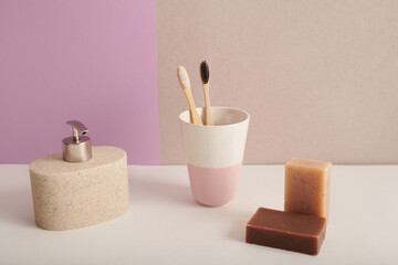 Still life shot of modern ceramic bathroom accessories with toothbrushes and soap bars against pink and pale brown background, copy space