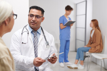 Middle-Eastern medical worker talking to senior woman while his female colleague calling for next patient in queue on background