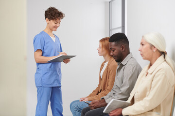 Obraz premium Group of three multi-ethnic people sitting in queue waiting for consultation in modern hospital, medical worker standing near checking attendance