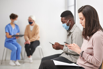 Obraz premium Horizontal shot of man and woman sitting on chair in hospital corridor waiting for consultation time surfing Internet on smartphone, doctor talking to mature woman on background