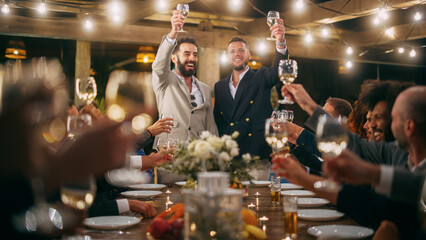Big Dinner Party with a Crowd of Multiethnic Diverse Friends Celebrating at a Restaurant. Handsome Happy Hosts Propose a Toast and Raise Wine Glasses while Sitting at a Table in the Evening.