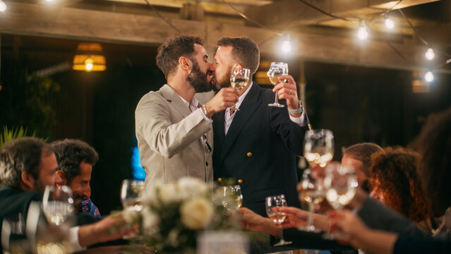 Big Dinner Party With A Crowd Of Multiethnic Diverse Friends Celebrating At A Restaurant. Handsome Happy Gay Hosts Propose A Toast And Raise Wine Glasses While Sitting At A Table In The Evening.