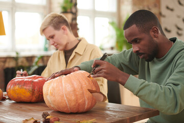 Focused young multi-ethnic people sitting at table and participating in pumpkin carving workshop...