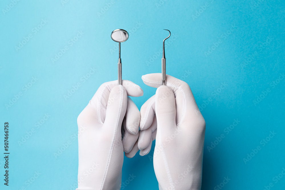 Wall mural The concept of oral health. A dentist in white gloves holds a double-sided periodontal probe and a stainless steel dental mirror on a turquoise background. Dental equipment close-up. Top view