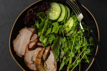 Hearty delicious lunch, fresh salad with cucumbers and microgreens of peas and with stewed pork, top view of the dark table with food