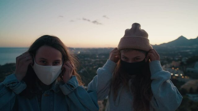 Attractive beautiful girls, young women, friends putting on protective safety medical masks, wearing casual clothing outside hugs each other on scenic nature background. Pandemic covid-19 disease time