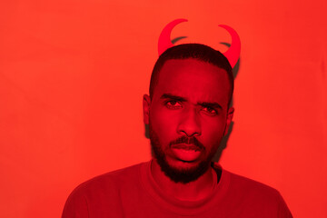 Portrait of frowning puzzled young African-American man in devil horns headband standing in red room, Halloween concept