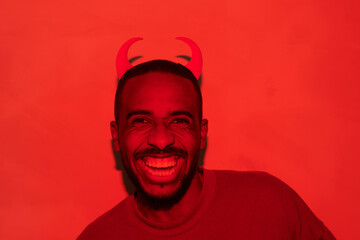 Portrait of ecstatic young Black devil guy with beard having crooked smile in red light, Halloween...