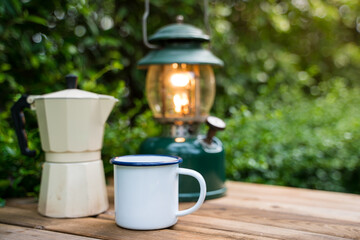 Selective focus white enamel coffee mug and coffee set in the garden with ancient lanterns in a camping atmosphere.