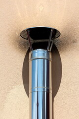 Shiny new metal flue pipe with wide cover on top held by four narrow metal strips next to stained family house wall with dirty smoke chimney stains on warm sunny spring day
