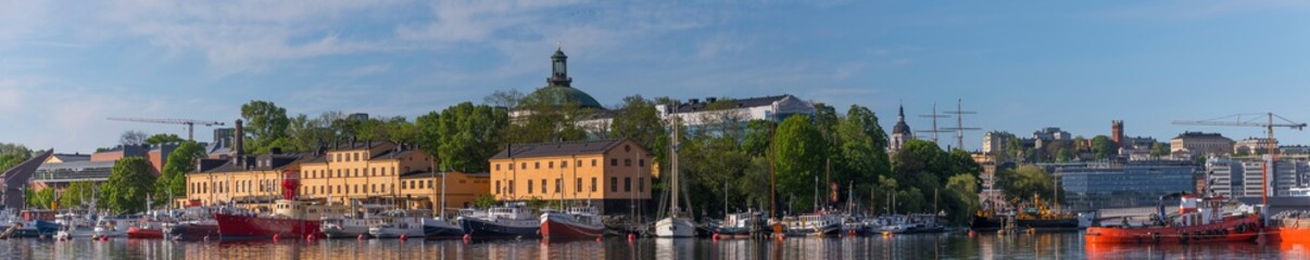Fototapeta na wymiar Panorama view over motor boats at piers in the islands Skeppsholmen and Blasieholmen, old military buildings and the district Södermalm in the background a sunny summer day in Stockholm