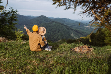Fototapeta na wymiar Back view of young mother in knit yellow beanie and cute little daughter sitting on mountain top near campfire, enjoying beautiful summer nature. Travel, adventure, wanderlust and tourism concept